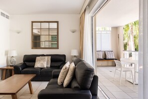 A modern living room with a black leather sofa, a wooden coffee table, and a large window with blinds. - Laguna 621 - Oasis on Hastings St (Noosa Heads)