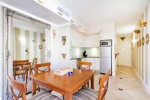 A dining area with a wooden table and chairs, a kitchen with white cabinets and a microwave, and a hallway with striped wallpaper and decorative wall sconces. - Laguna 622 - Nautical Romance in Noosa (Noosa Heads)