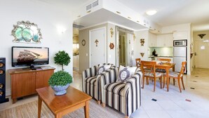 Ein Wohnzimmer mit Fernseher, Sofa, Esstisch, Stühlen und einer Küche im Hintergrund.