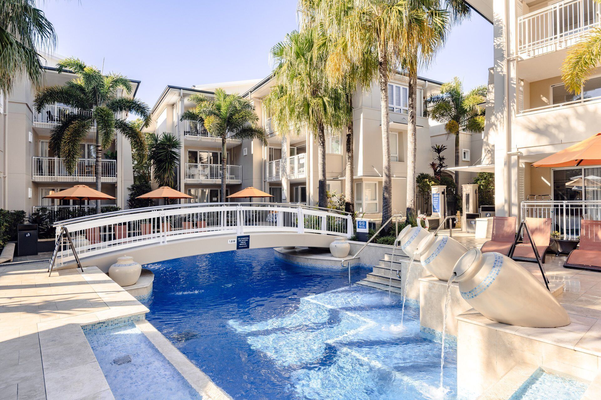 A swimming pool with a bridge, surrounded by palm trees and a multi-story building with balconies.