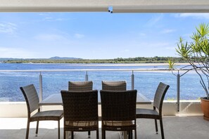 A terrace with wicker chairs and a table overlooking a beach and water.