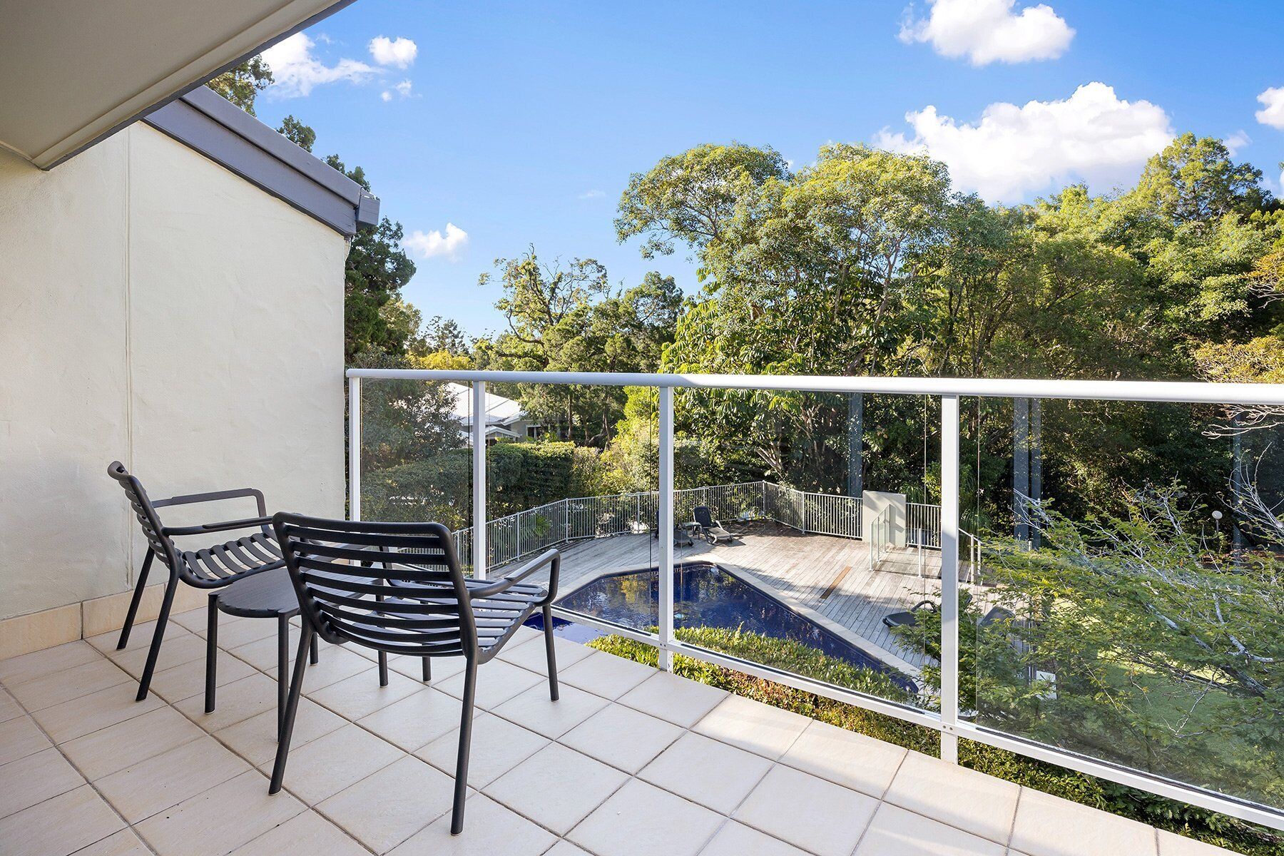 Un balcón con dos sillas y barandilla de vidrio con vista a un jardín y un puente.