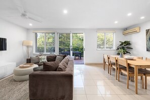 A modern living room with a sofa, a dining table with chairs, and a large window with blinds.
