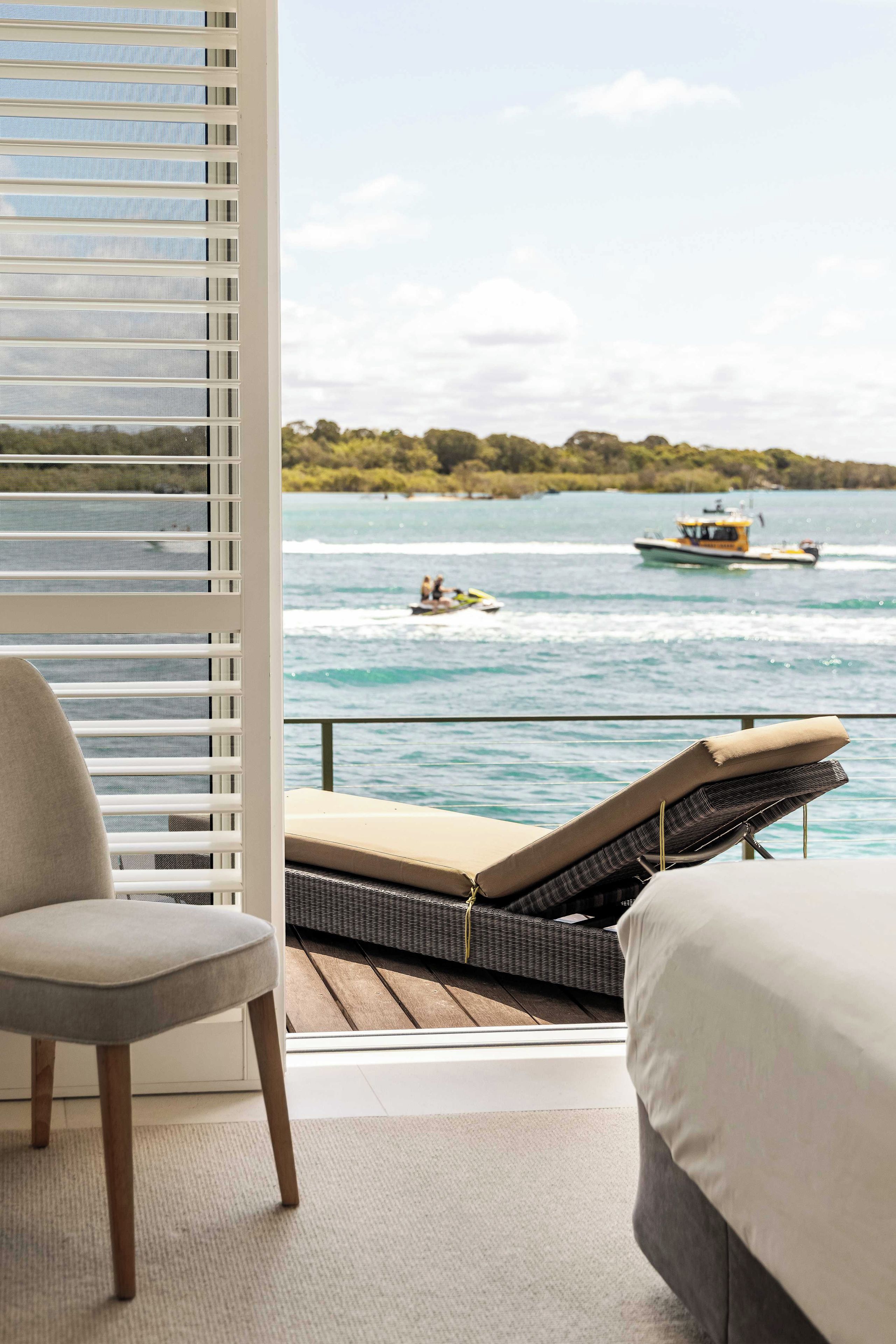 A room with a view of a body of water, a boat, and a lounge area with a chair and a bed.