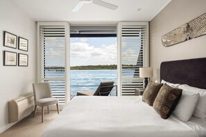 Un dormitorio con una cama grande, una silla, un escritorio y vistas a un cuerpo de agua a través de contraventanas abiertas.