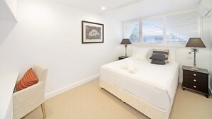 Een slaapkamer met een bed, twee nachtkastjes, een stoel, een raam met zonwering en een lijst met een afbeelding aan de muur.