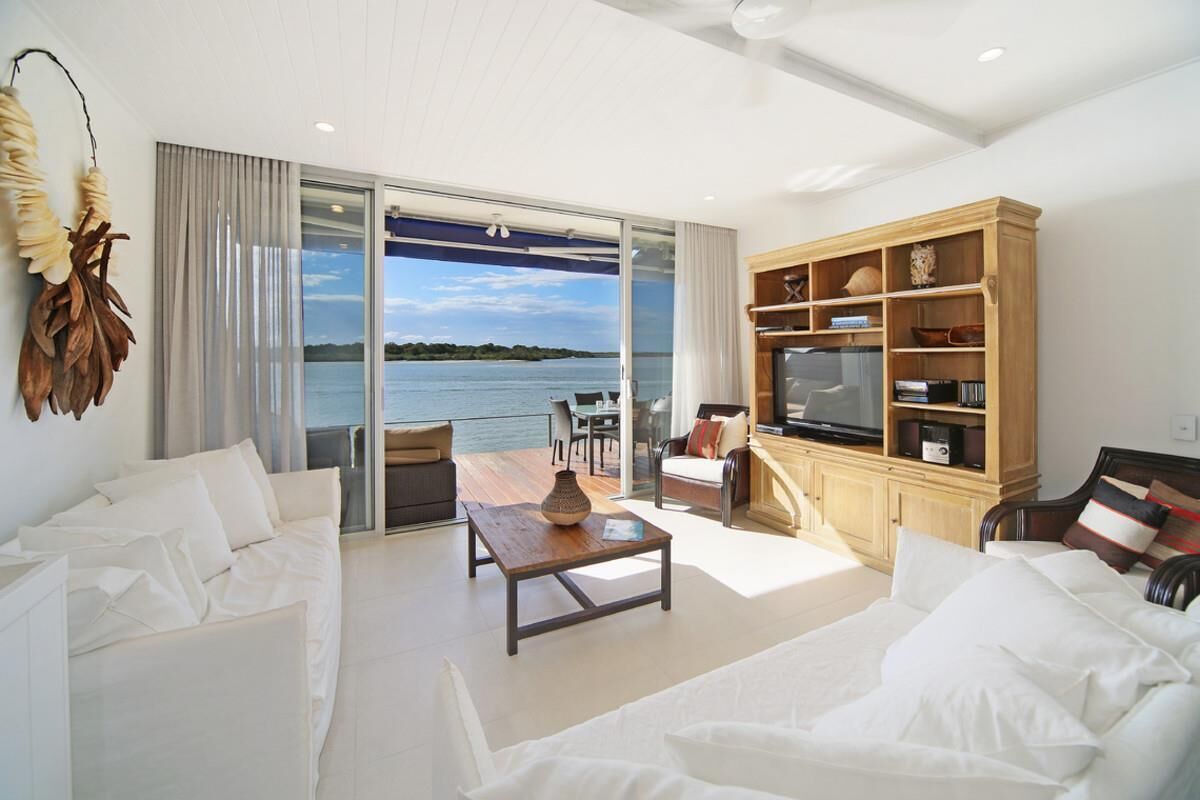 Una sala de estar moderna con un amplio ventanal corredero que da a un balcón con vistas al agua. Hay un sofá, una mesa de centro y un mueble para la televisión con estantes.