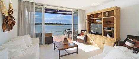 Ein modernes Wohnzimmer mit einem großen Schiebeglasfenster, das auf einen Balkon mit Blick auf das Wasser führt. Es gibt ein Sofa, einen Couchtisch und eine TV-Kommode mit Regalen.