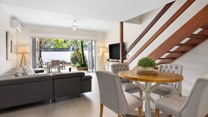 Een moderne woonkamer met een grijze bank, een ronde houten eettafel met stoelen, een televisie en een trap met houten leuningen.