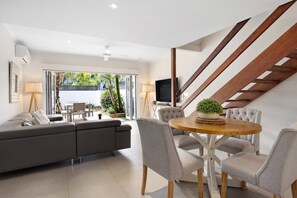 Ein modernes Wohnzimmer mit einem grauen Sofa, einem runden Holzesstisch mit Stühlen, einem Fernseher und einer Treppe mit Holzwangen.