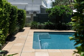A swimming pool with a tiled deck, surrounded by greenery and a building in the background. - Noosa Court 4 - Beachfront Bliss (Noosa Heads)