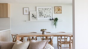 A dining area with a wooden table and chairs, a white wall with artwork, and a plant.