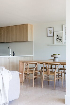 A modern kitchen with wooden cabinets, a white countertop, and a dining area with a wooden table and chairs. - Noosa Court 4 - Beachfront Bliss (Noosa Heads)