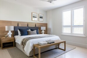 A bedroom with a bed, two bedside tables with lamps, a window with blinds, and a bedside table with books.