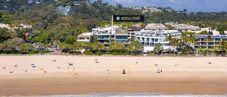 A beachfront hotel named Noosa Court with a sandy beach, ocean view, and a hilly backdrop.