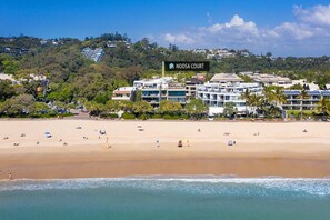 A beachfront hotel named Noosa Court with a sandy beach, ocean view, and a hilly backdrop.