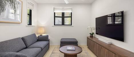 Una sala de estar moderna con un sofá gris, una mesa de centro redonda, un ventilador de techo, un televisor montado en la pared y una ventana con persianas.