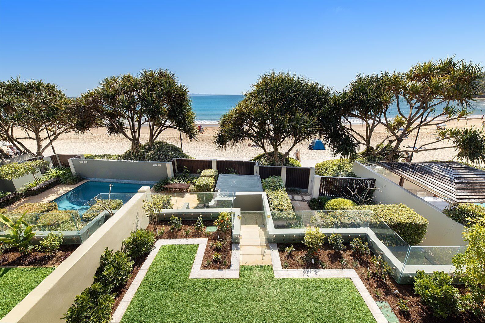 A pool area with a view of the beach, surrounded by greenery and a clear sky.