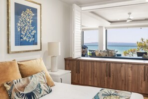 Een slaapkamer met een groot raam, een bed met kussens, een nachtkastje en een ingelijst kunstwerk.