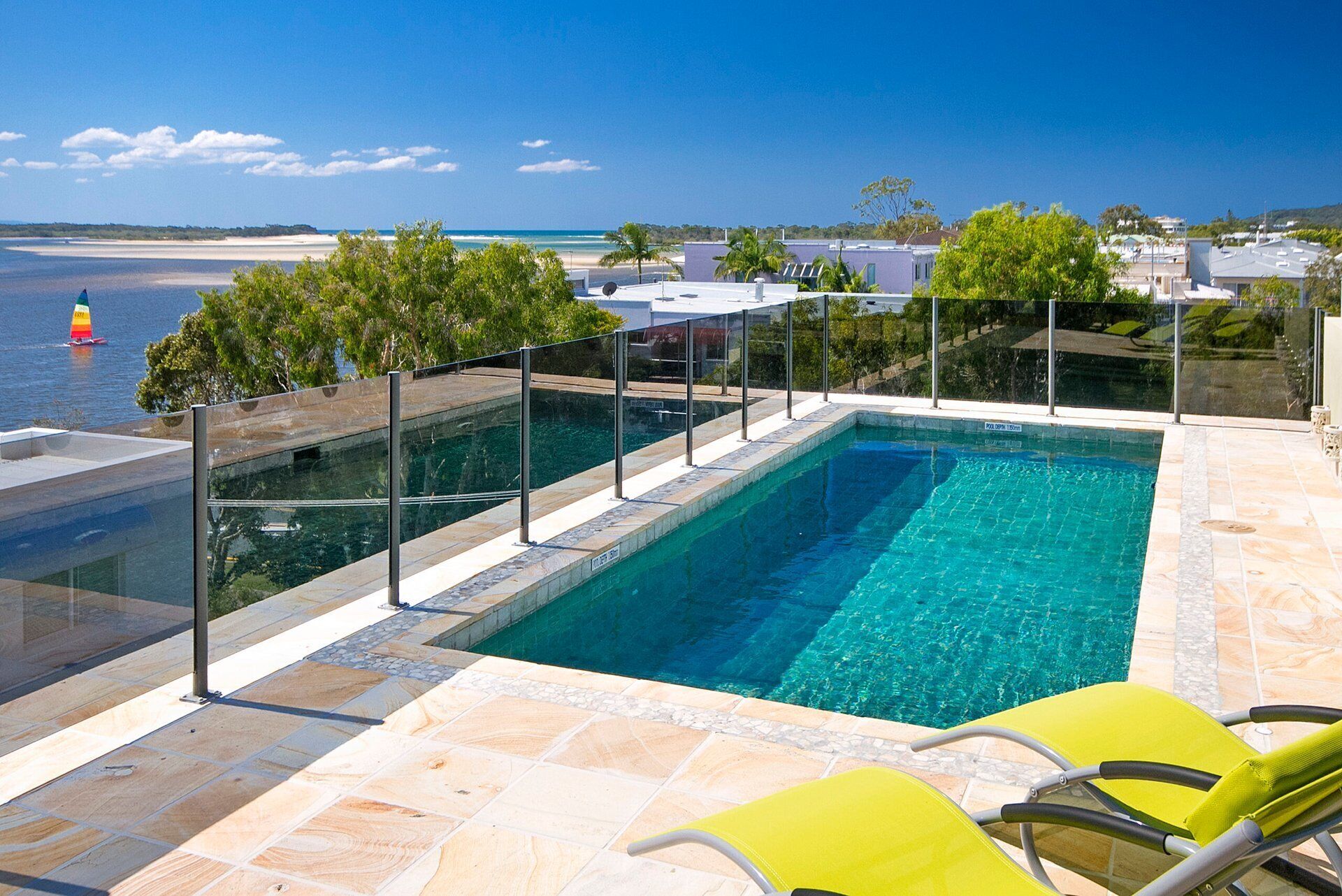 Une piscine séparée du reste par une paroi en verre, deux chaises longues jaunes et une vue sur l’océan et la plage.