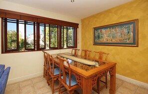 A dining area with a wooden table and chairs, large windows with blinds, and a painting on the wall.