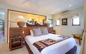 A hotel room with a large bed, a wooden headboard, a bedside table, a chair, a mirror, and a ceiling fan. - Noosa Dua 2 - Balinese Retreat (Noosaville)