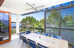 A dining area with a table and chairs, a ceiling fan, and a view of the sea through large windows. - Noosa Dua 2 - Balinese Retreat (Noosaville)