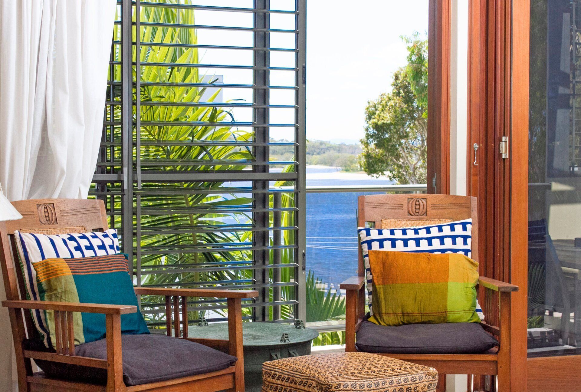 Two wooden chairs with cushions, a small table, and a view of greenery and a body of water through a window with a metal grid.
