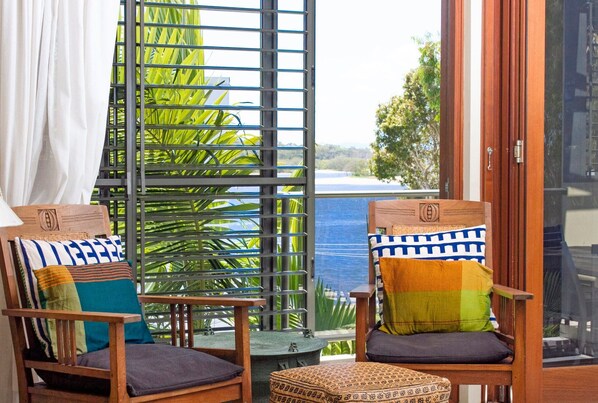Two wooden chairs with cushions, a small table, and a view of greenery and a body of water through a window with a metal grid.