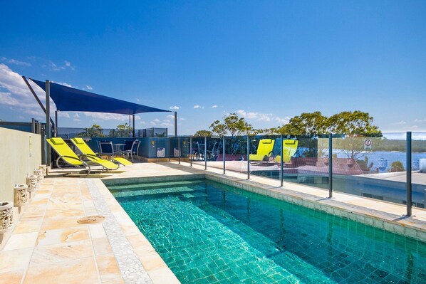 A swimming pool with clear blue water, surrounded by lounge chairs and a shaded area.