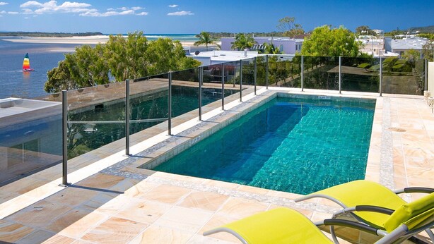 A swimming pool with a glass barrier, two yellow lounge chairs, and a view of the ocean and beach.