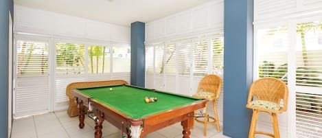 A pool table with a net, surrounded by wicker chairs, in a room with white shutters and a tiled floor.