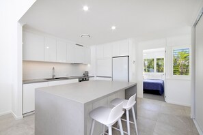Una cucina moderna con mobili bianchi, un'isola centrale con sgabelli da bar e vista su una camera da letto attraverso una porta aperta.
