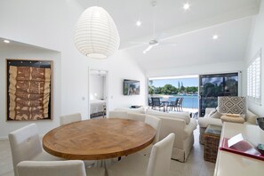 A modern living room with a round wooden table, white sofas, a ceiling fan, and a large window overlooking a pool.