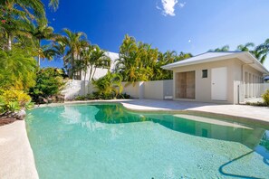 A clear swimming pool with a white deck, surrounded by tropical vegetation and a building with a flat roof.