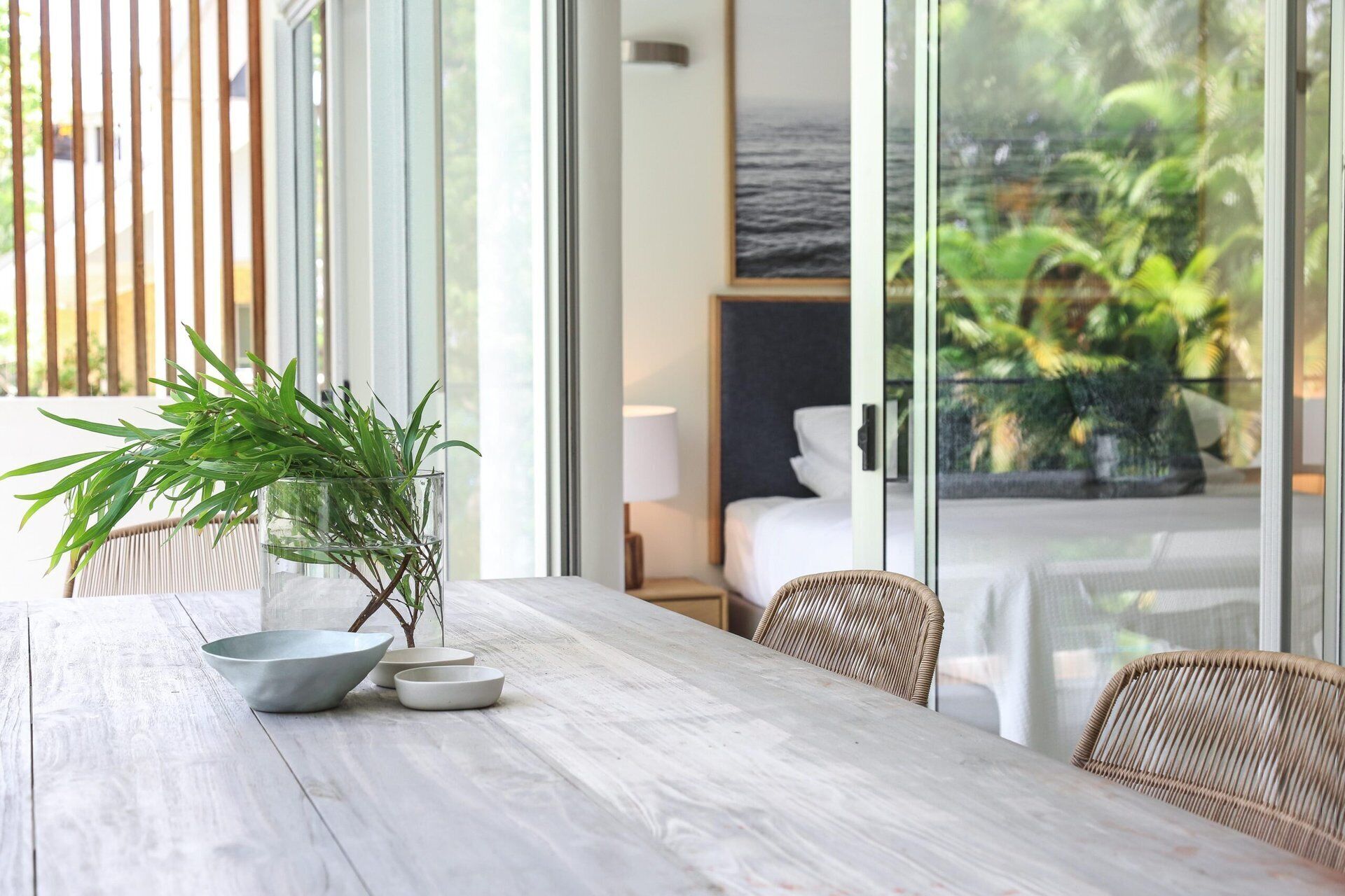 A wooden table with a potted plant and bowls, a wicker chair, and a view of a bedroom with a bed and a nightstand.