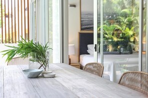 A wooden table with a potted plant and bowls, a wicker chair, and a view of a bedroom with a bed and a nightstand.