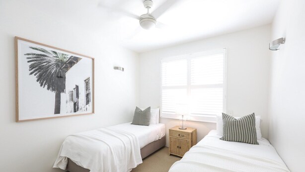 Un dormitorio con dos camas individuales, un ventilador de techo, una ventana con persianas y un cuadro en la pared.