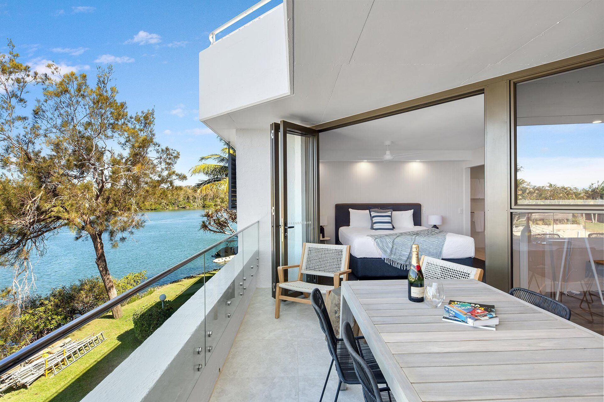 A modern hotel room with a balcony overlooking a body of water, a bed with a view, and a dining area with a table and chairs.