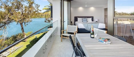 A modern hotel room with a balcony overlooking a body of water, a bed with a view, and a dining area with a table and chairs.
