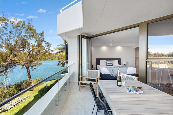 A modern hotel room with a balcony overlooking a body of water, a bed with a view, and a dining area with a table and chairs.