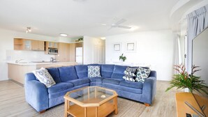 Een moderne woonkamer met een blauwe modulaire bank, een glazen salontafel en een keuken op de achtergrond.
