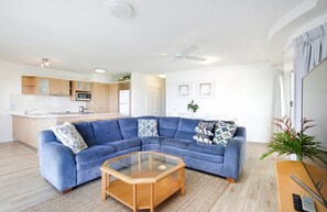 A modern living room with a blue sectional sofa, a glass coffee table, and a kitchen in the background. - Nature's Haven Coastal Escape (Noosa Heads)