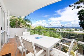 Varanda com mesa e cadeiras brancas, vista para um horizonte urbano com palmeiras e céu claro.