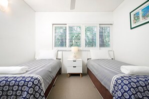 Quarto com duas camas, mesa de cabeceira, abajur e janela com persianas.