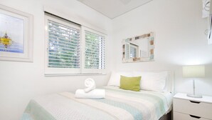 Een slaapkamer met een bed, een nachtkastje, een raam met zonwering, een lijst met foto en een spiegel.