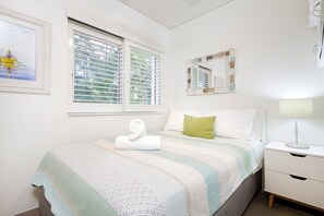 A bedroom with a bed, a bedside table, a window with blinds, a framed picture, and a mirror. - Nature's Haven Coastal Escape (Noosa Heads)