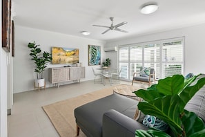 Un salon moderne comprenant un canapé, une table à manger avec des chaises, un ventilateur de plafond et de grandes fenêtres équipées de stores.