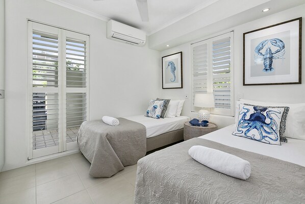Two single beds with neatly made beds and matching pillows, a bedside table with a lamp and decorative items, and framed artworks adorn this stylish room.