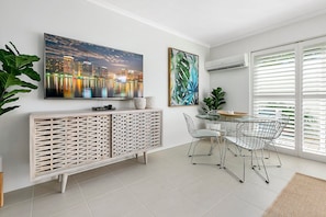 Un espace repas moderne avec une table en verre et des chaises en métal, une console blanche agrémentée d’un paravent décoratif, et deux œuvres d’art encadrées accrochées au mur.